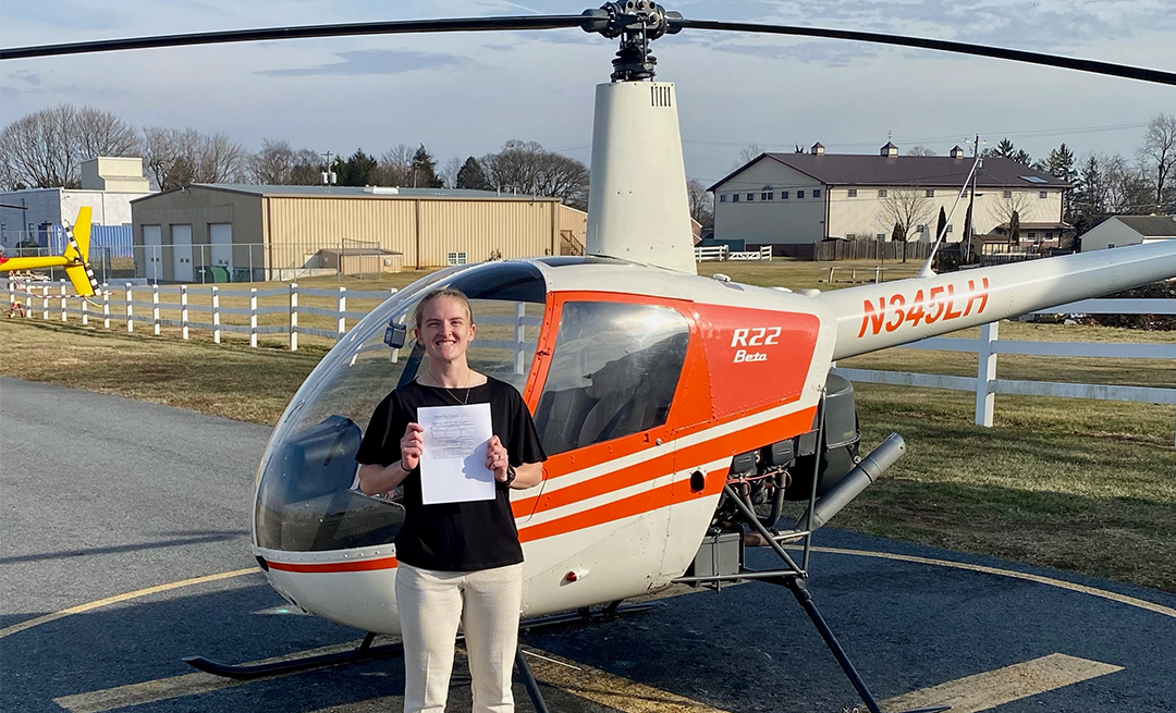 Person in light pants and dark shirt poses in front of orange and white helicopter on airfield